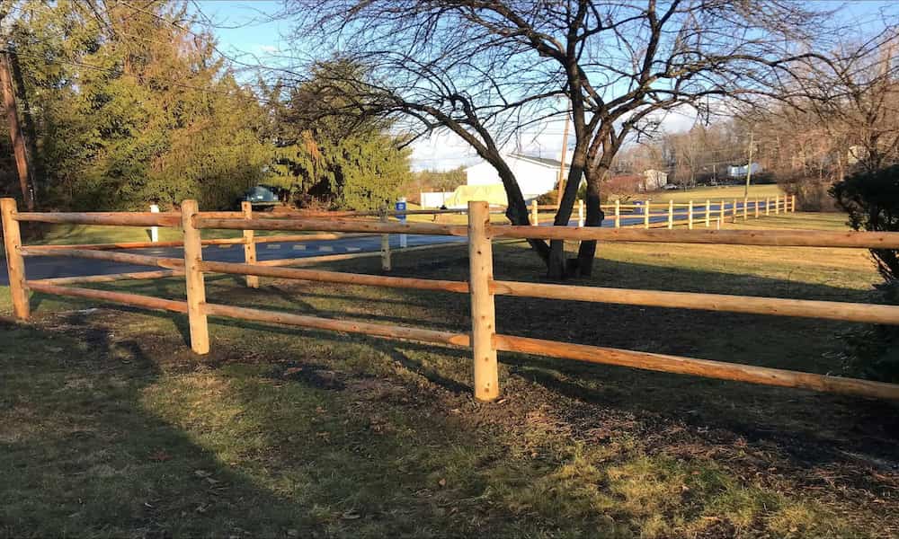 A wooden fence runs alongside a quiet rural road, bordered by bare trees and grass.