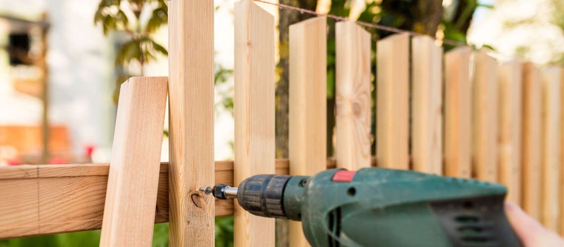 A close-up of a person using a green cordless power drill to screw a wooden picket
